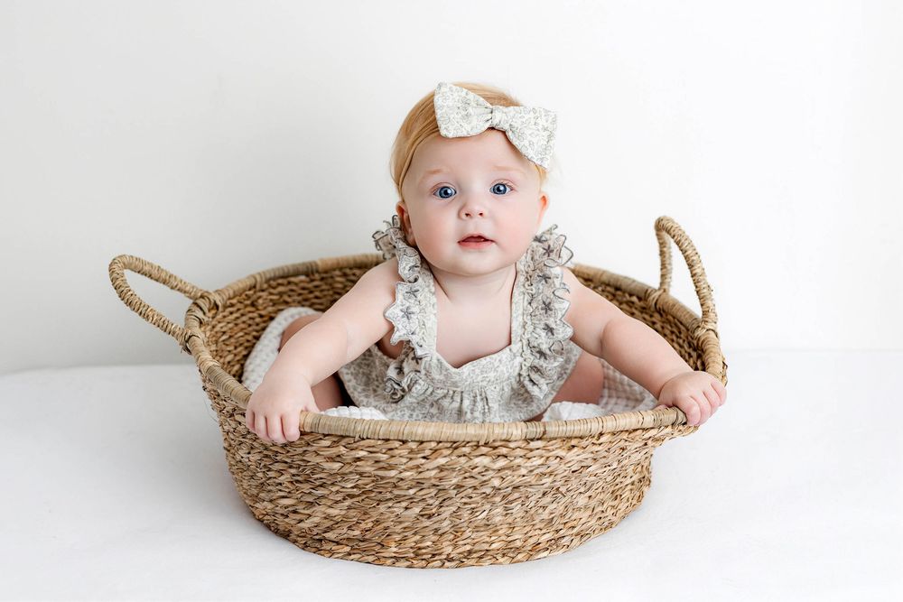 Adorable baby girl in a floral romper and bow headband sitting in a woven basket on a white background.