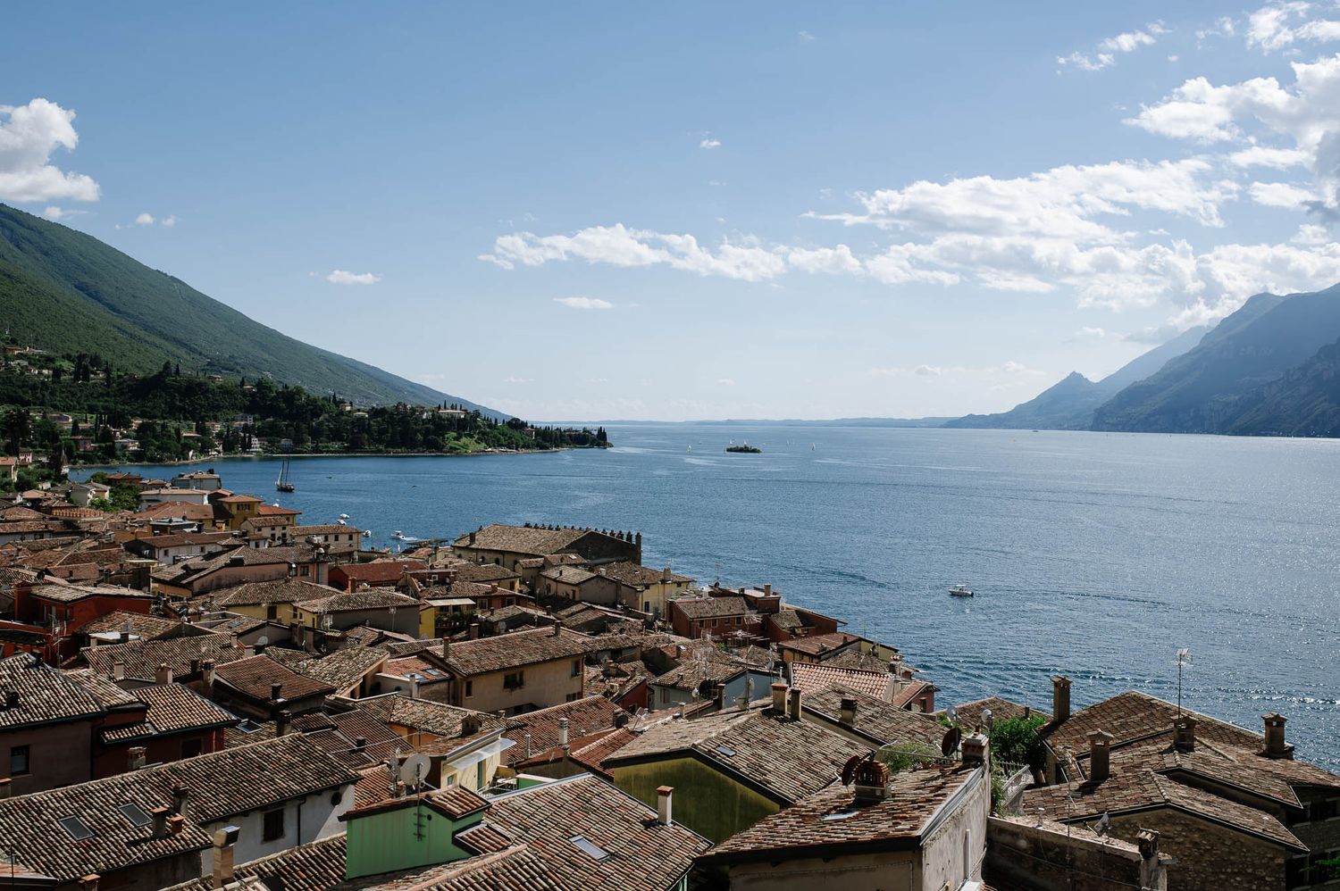 Aerial view of a picturesque Italian lakeside town with terracotta rooftops, mountains, and blue waters under a partly cloudy sky.