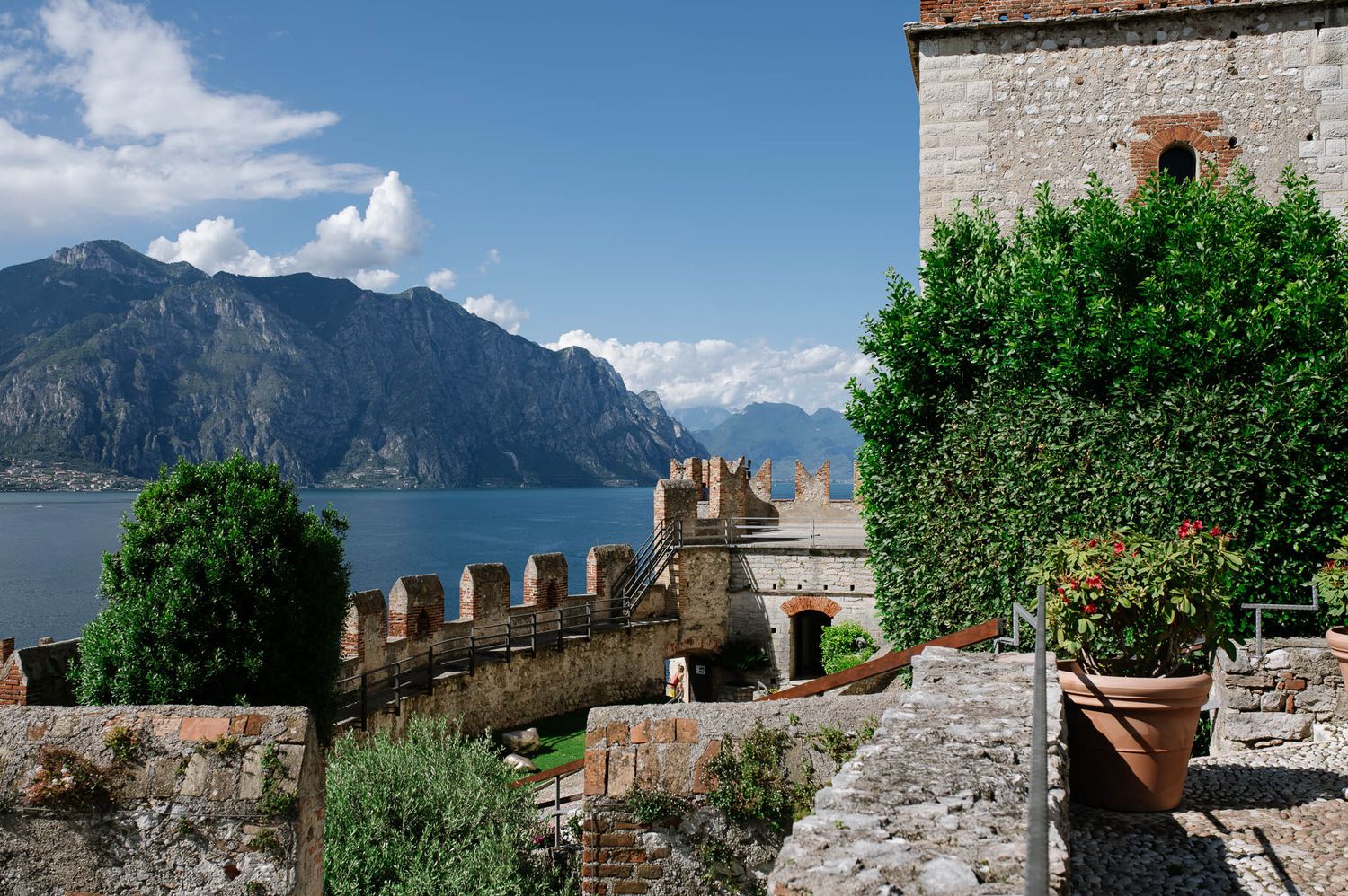 Medieval castle with battlements overlooking a serene alpine lake surrounded by mountains under a blue sky.