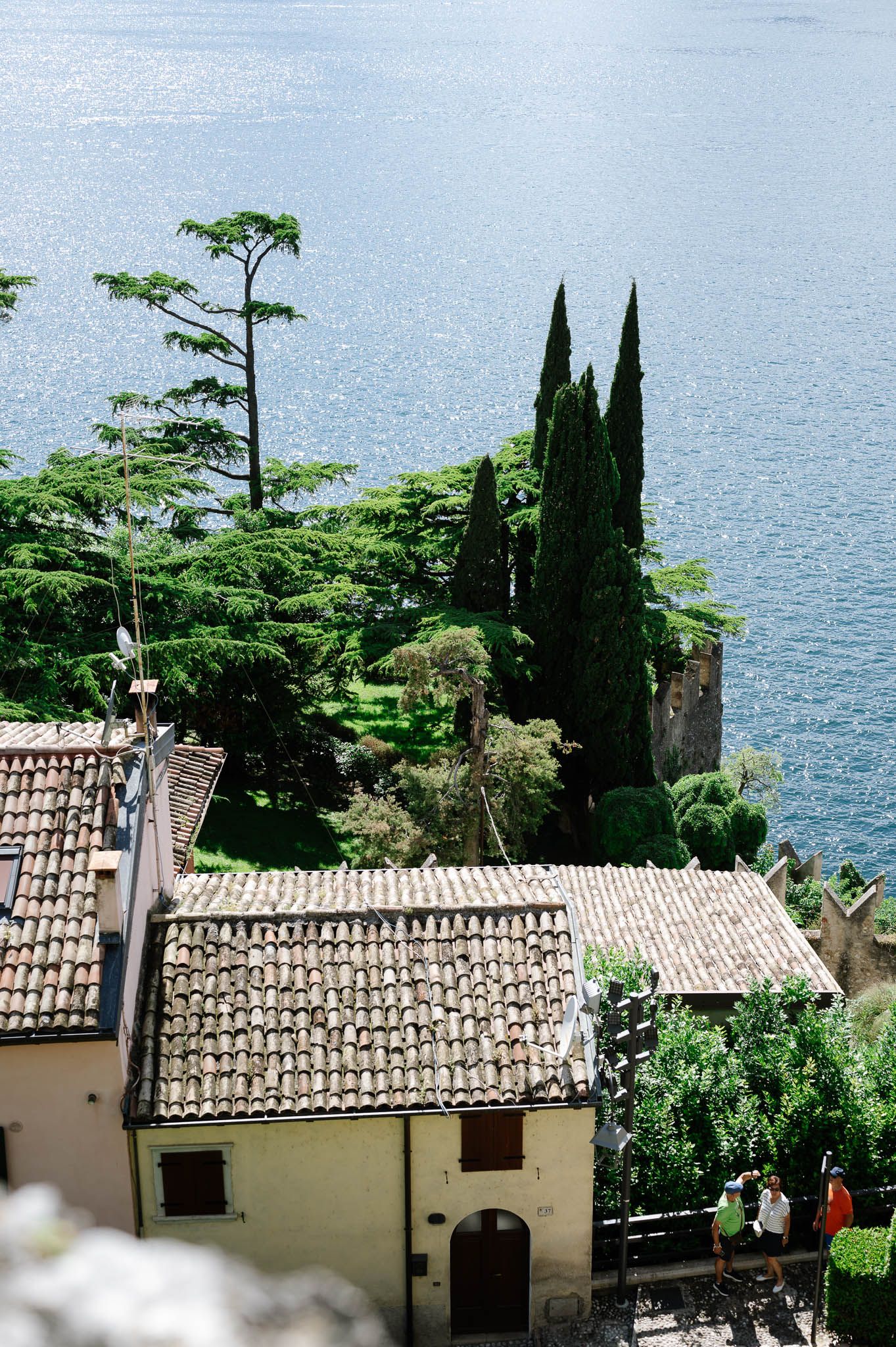 Aerial view of Italian lakeside village with terracotta rooftops, cypress trees, and shimmering blue lake in background.
