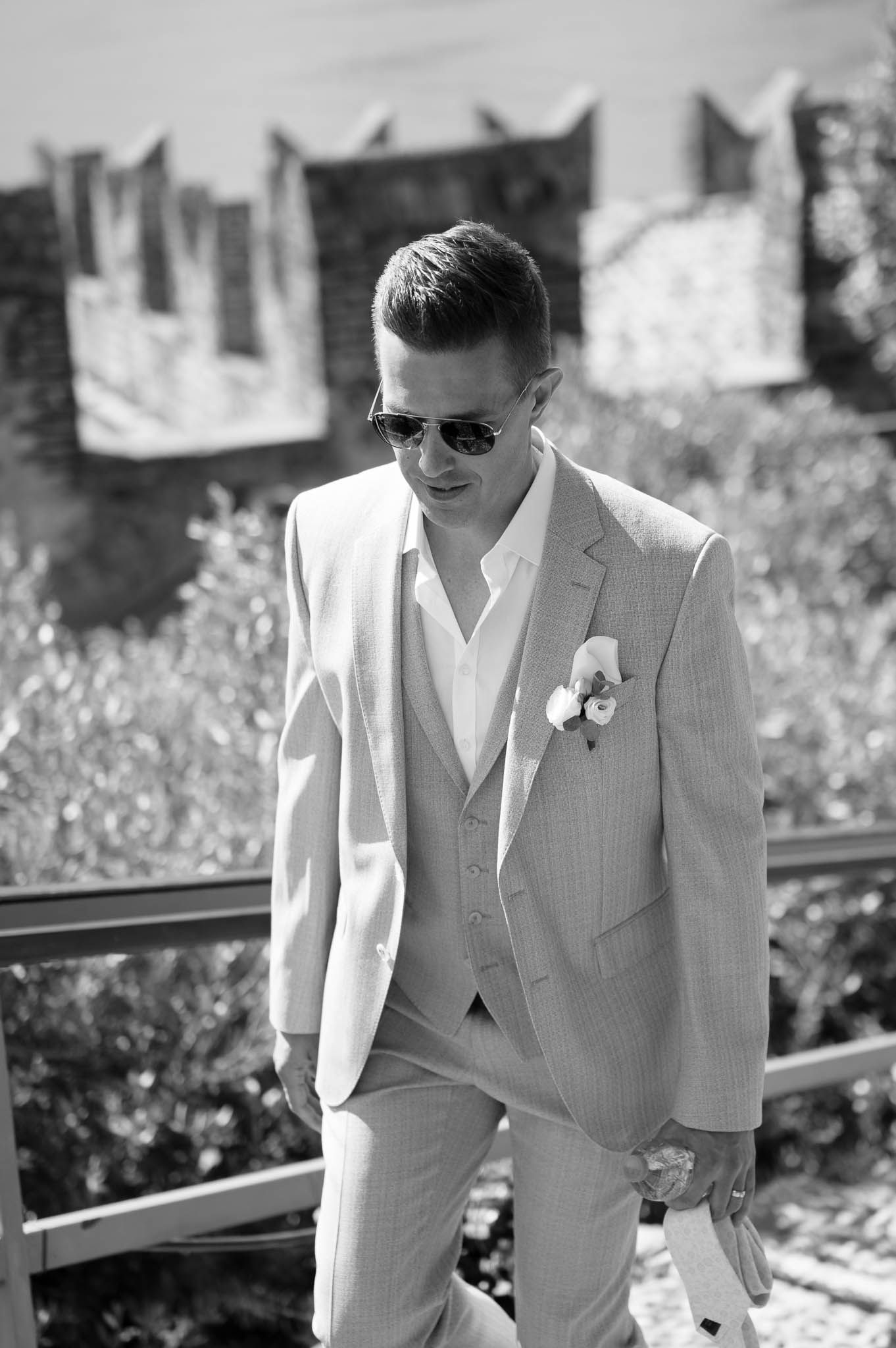 Elegant groom in light suit with boutonniere and sunglasses posing near castle battlements on wedding day.