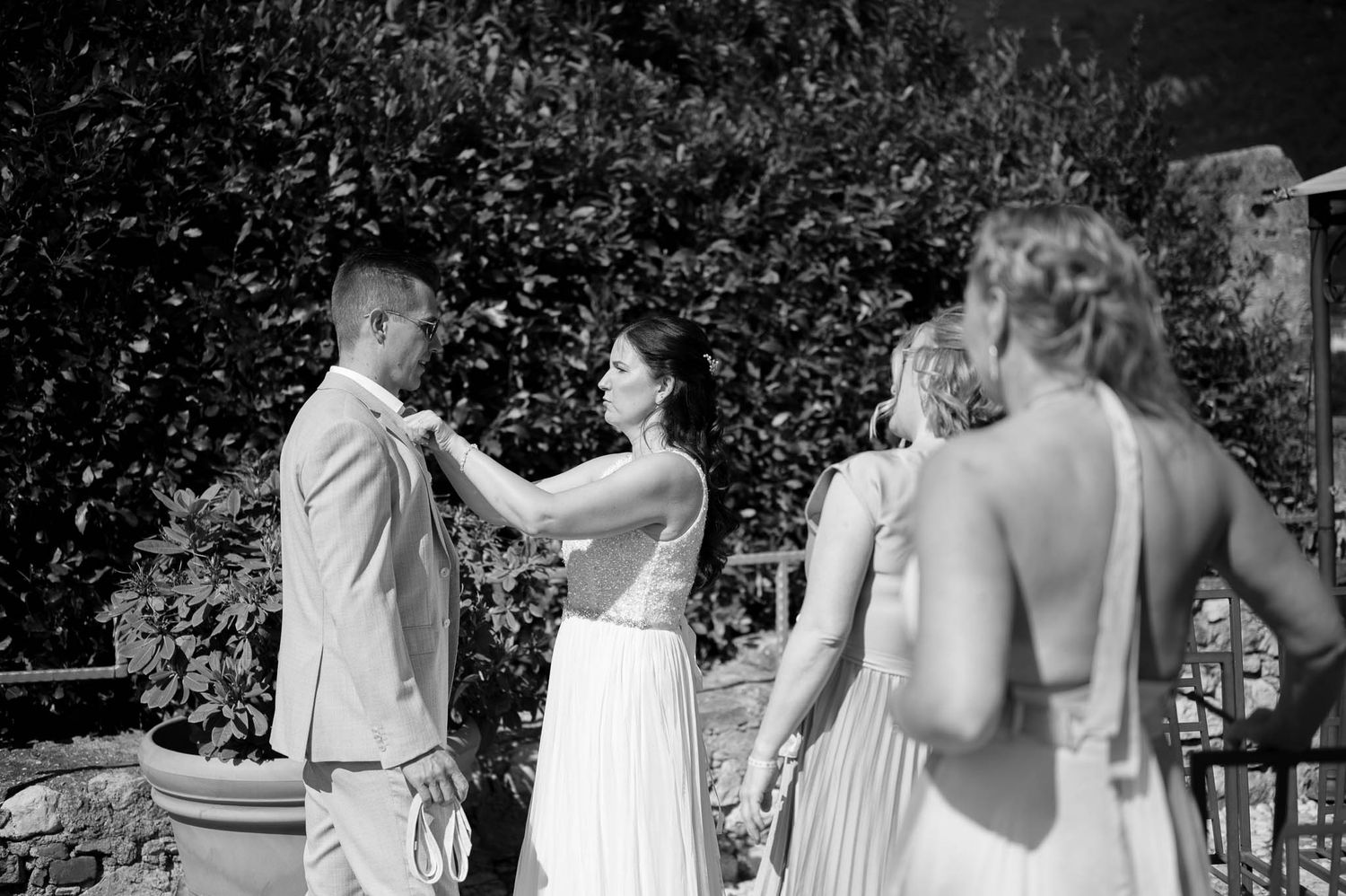 Bride adjusts groom's suit lapel as bridesmaids stand nearby in a black and white outdoor wedding photo.