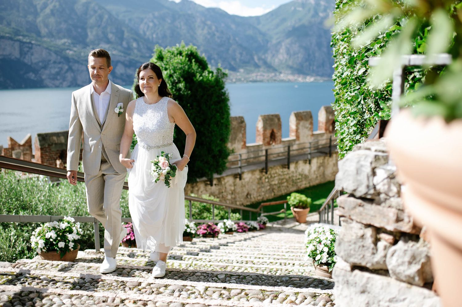 Bride and groom walk hand-in-hand down sunlit stone steps at Italian castle wedding venue overlooking lake.
