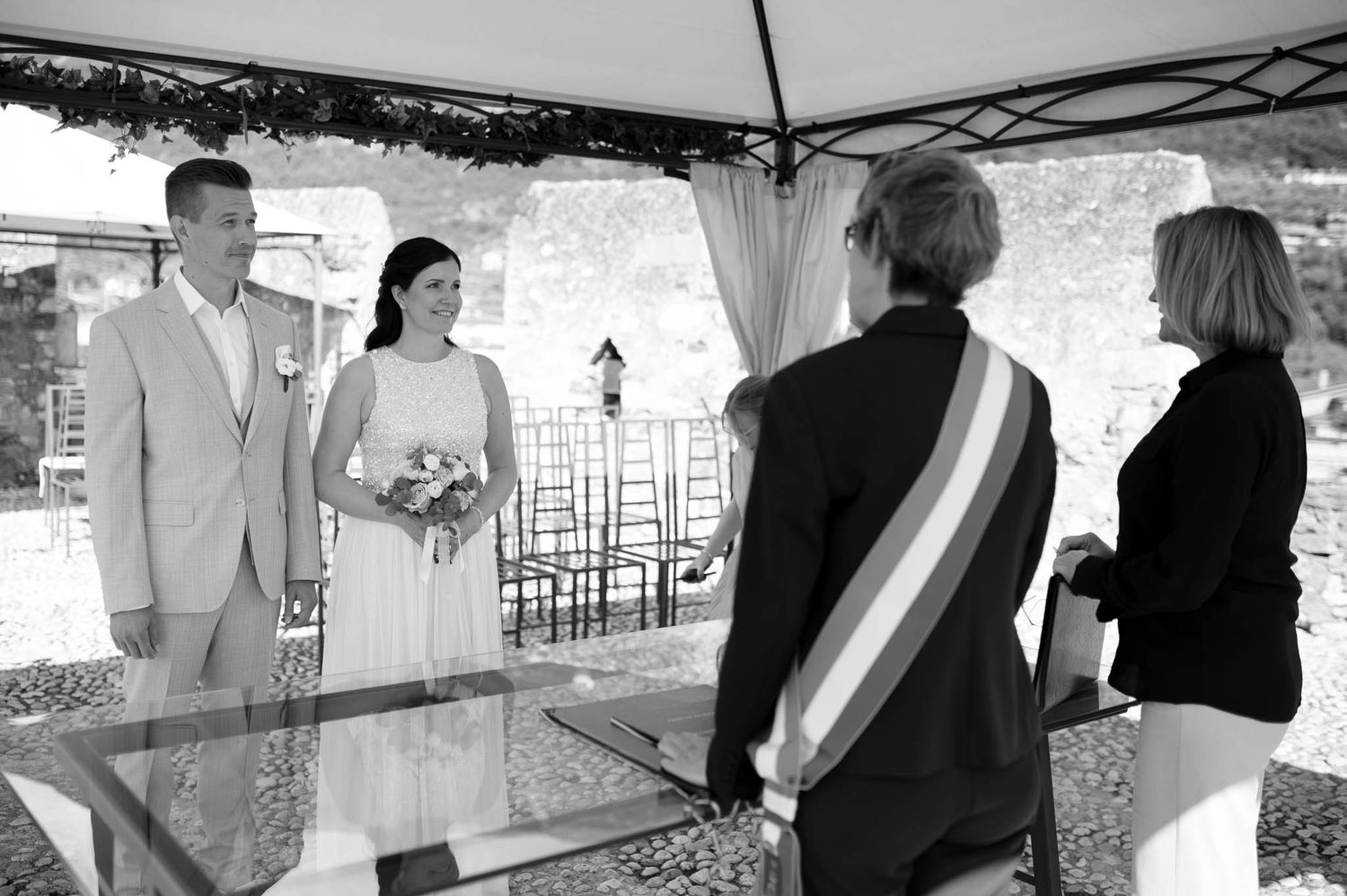 Black and white photo of a wedding ceremony under a canopy with bride, groom, and officiant.