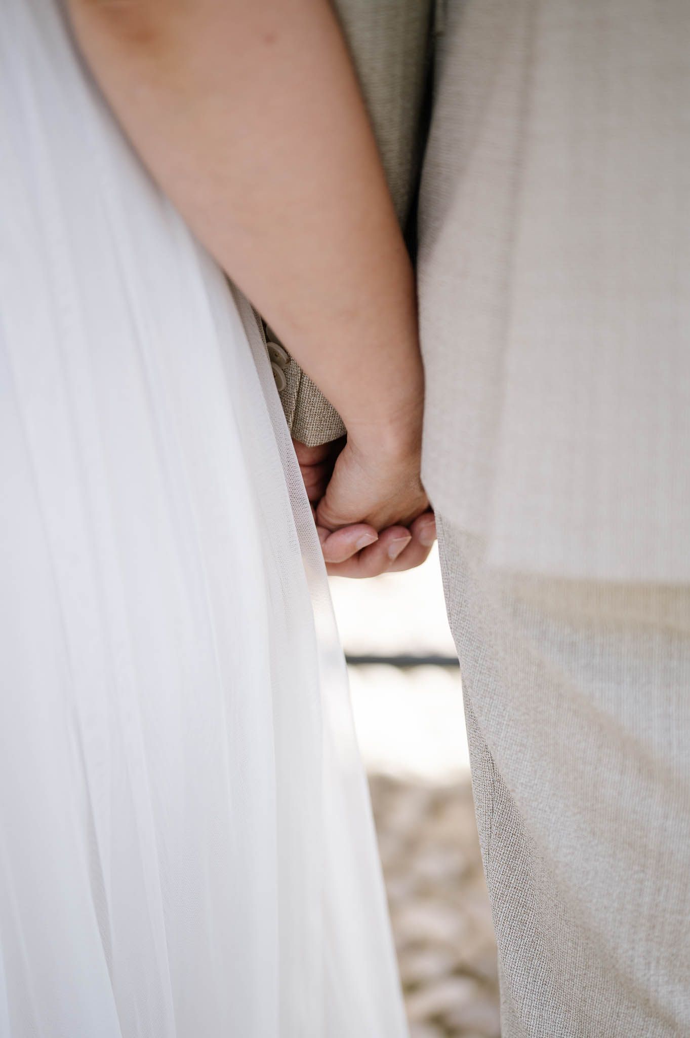 Close-up of bride and groom holding hands, white wedding dress and beige suit visible.