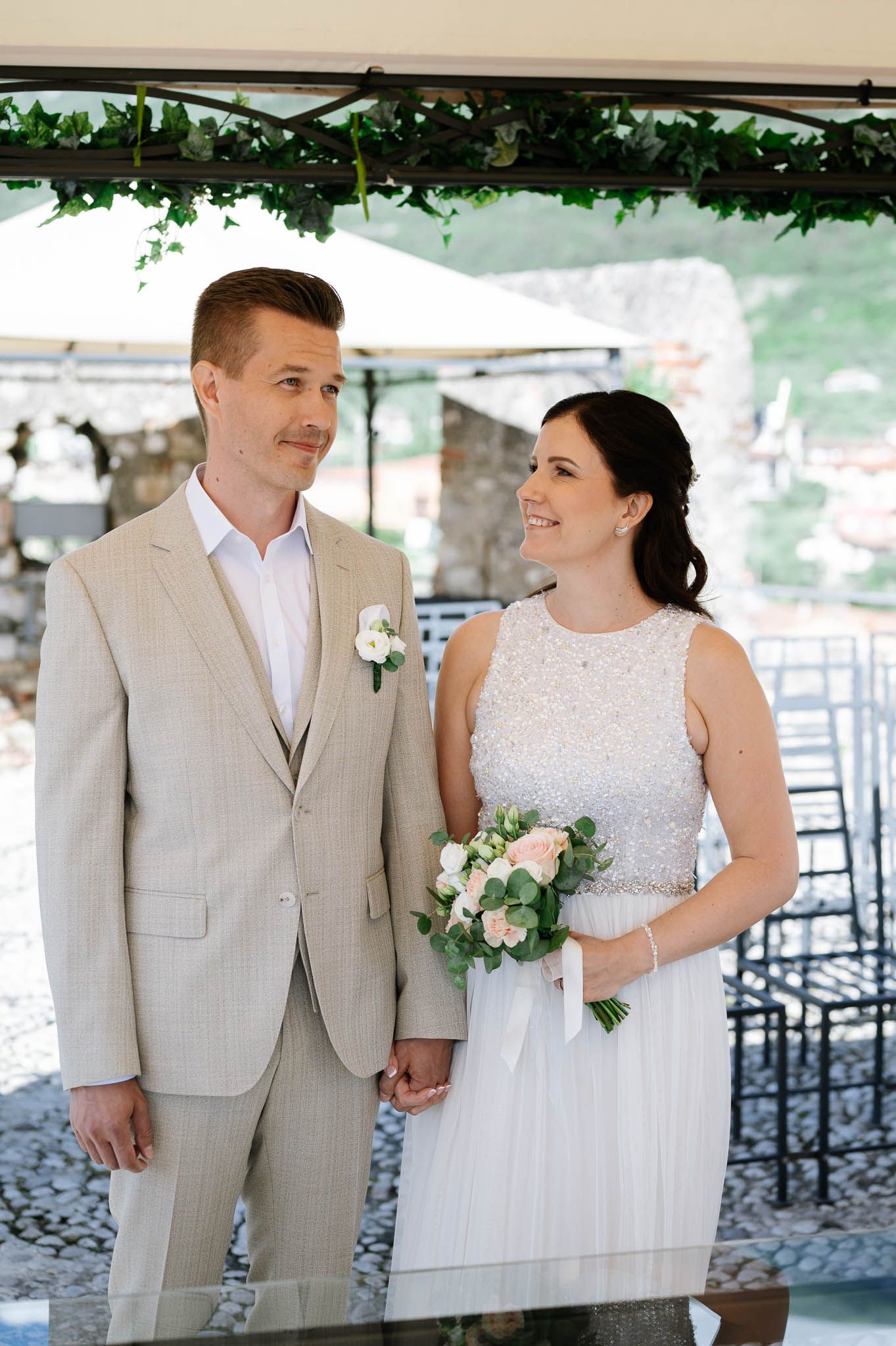 Bride and groom holding hands during outdoor wedding ceremony, bride in beaded gown holding pink bouquet, groom in tan suit.