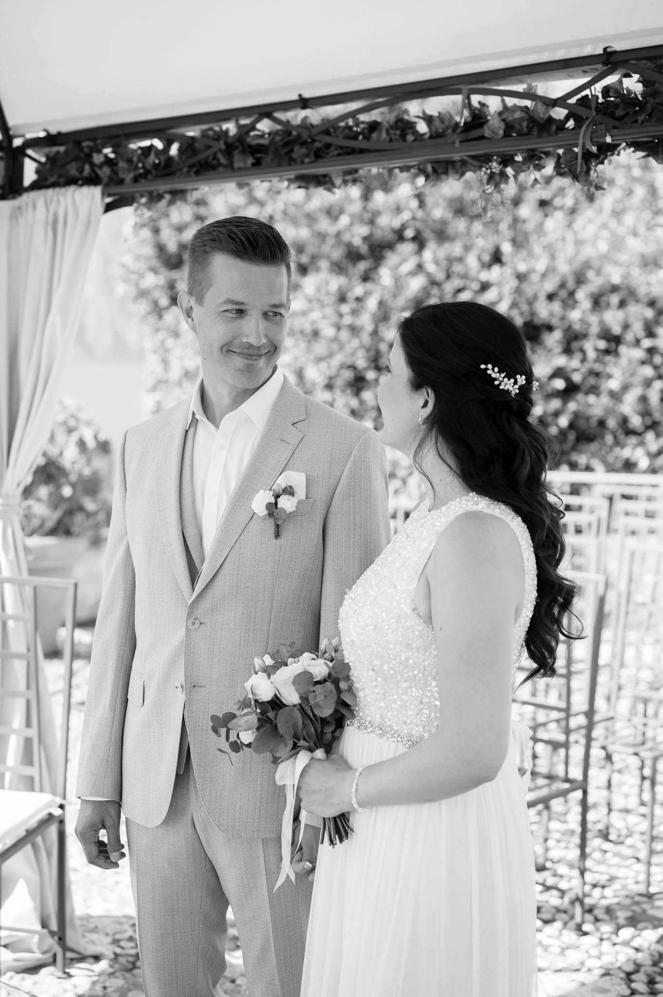 Black and white portrait of smiling groom in tan suit and bride holding bouquet during outdoor wedding ceremony.