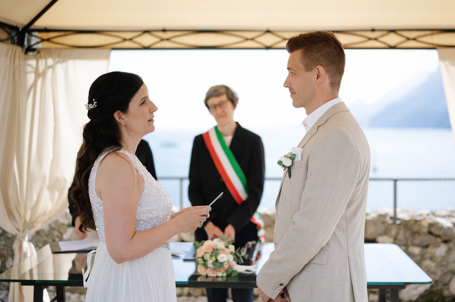 Bride reading vows to groom at outdoor lakeside wedding ceremony with officiant wearing Italian sash in background.