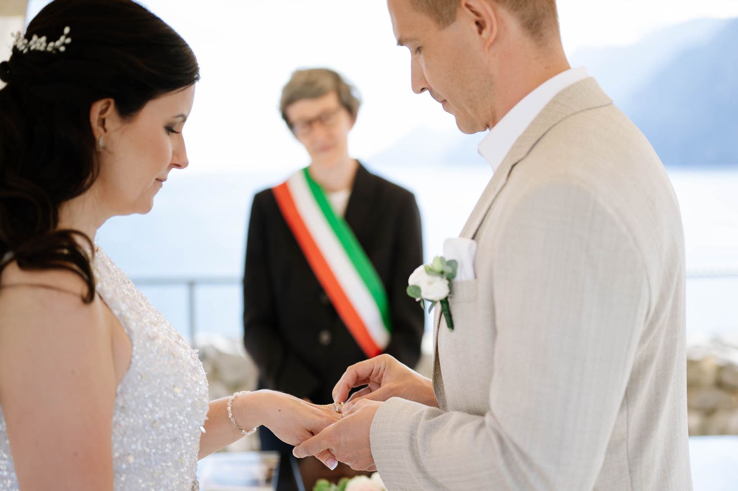 Groom places wedding ring on brides finger during intimate lakeside ceremony officiated by sash-wearing official.