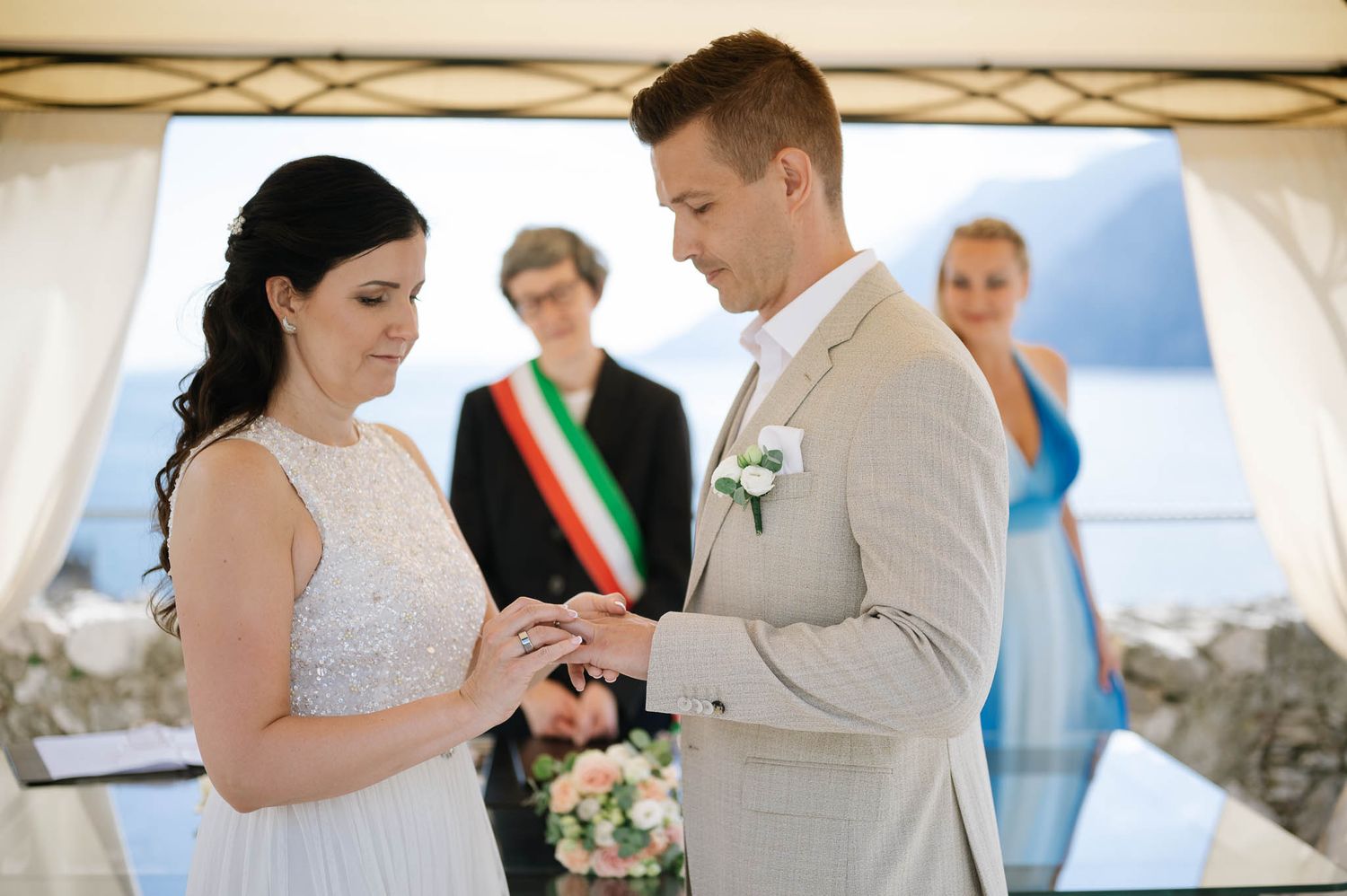 Bride placing wedding ring on grooms finger during outdoor lakeside ceremony in Italy.