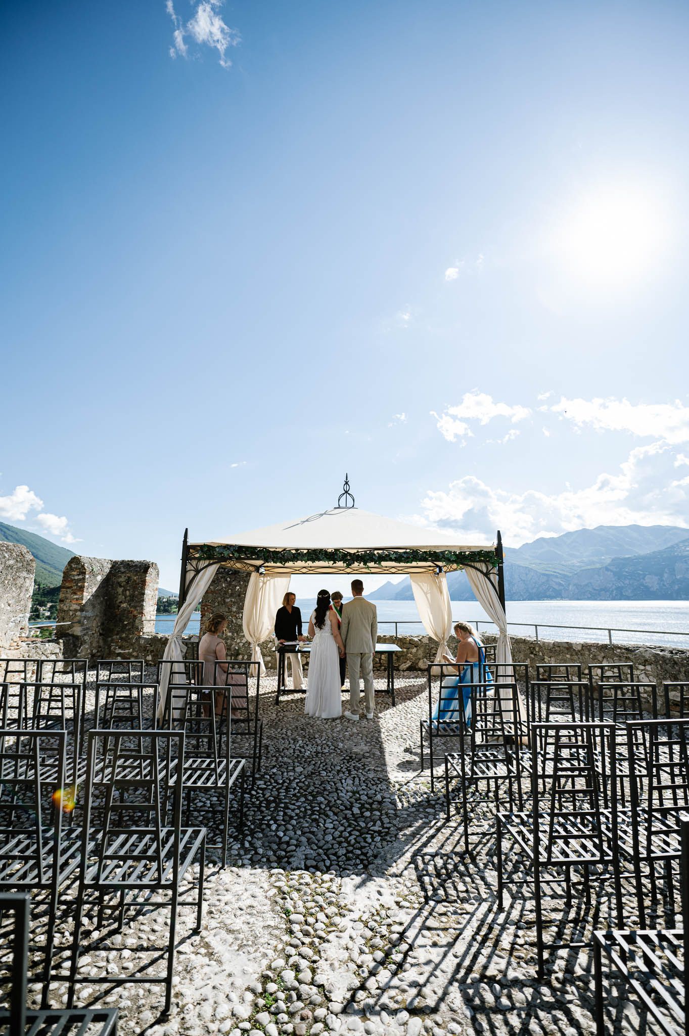 Outdoor wedding ceremony under elegant gazebo with lake and mountain backdrop on sunny day.