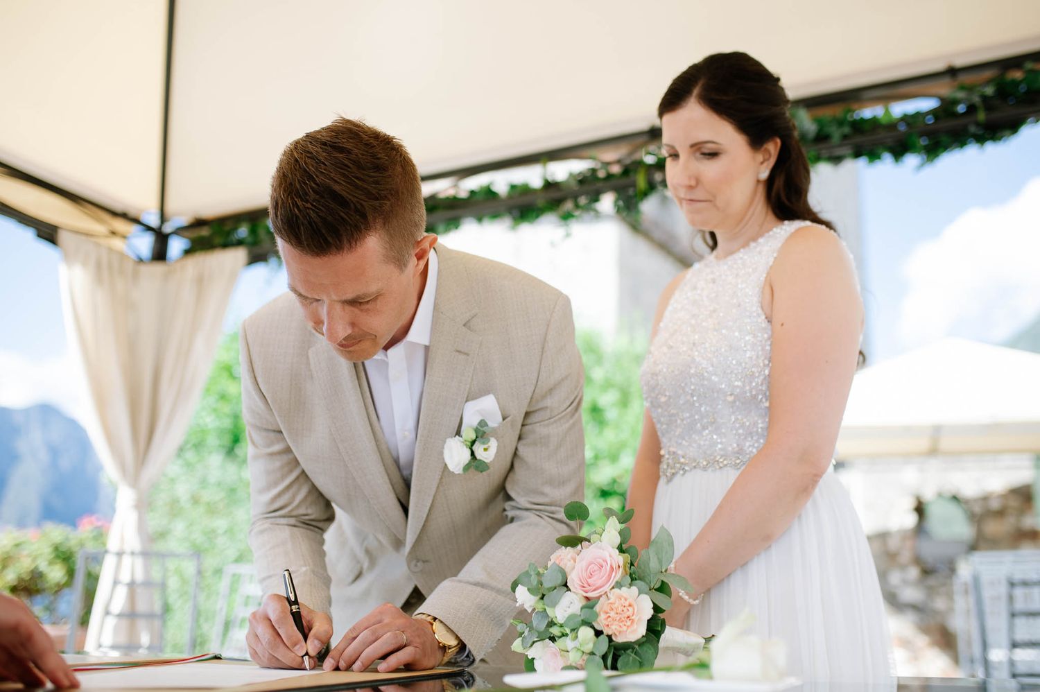 Groom signing wedding register while bride holds pink floral bouquet at outdoor ceremony.