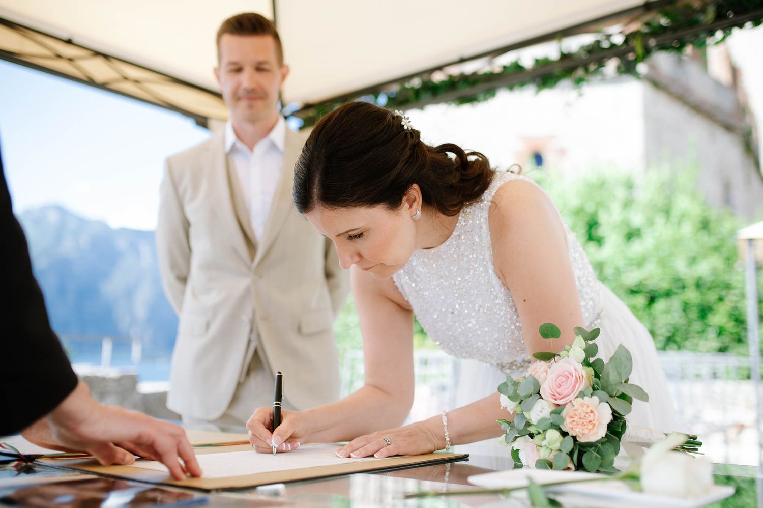 Bride signs wedding register at outdoor ceremony while groom watches, holding pink rose bouquet.