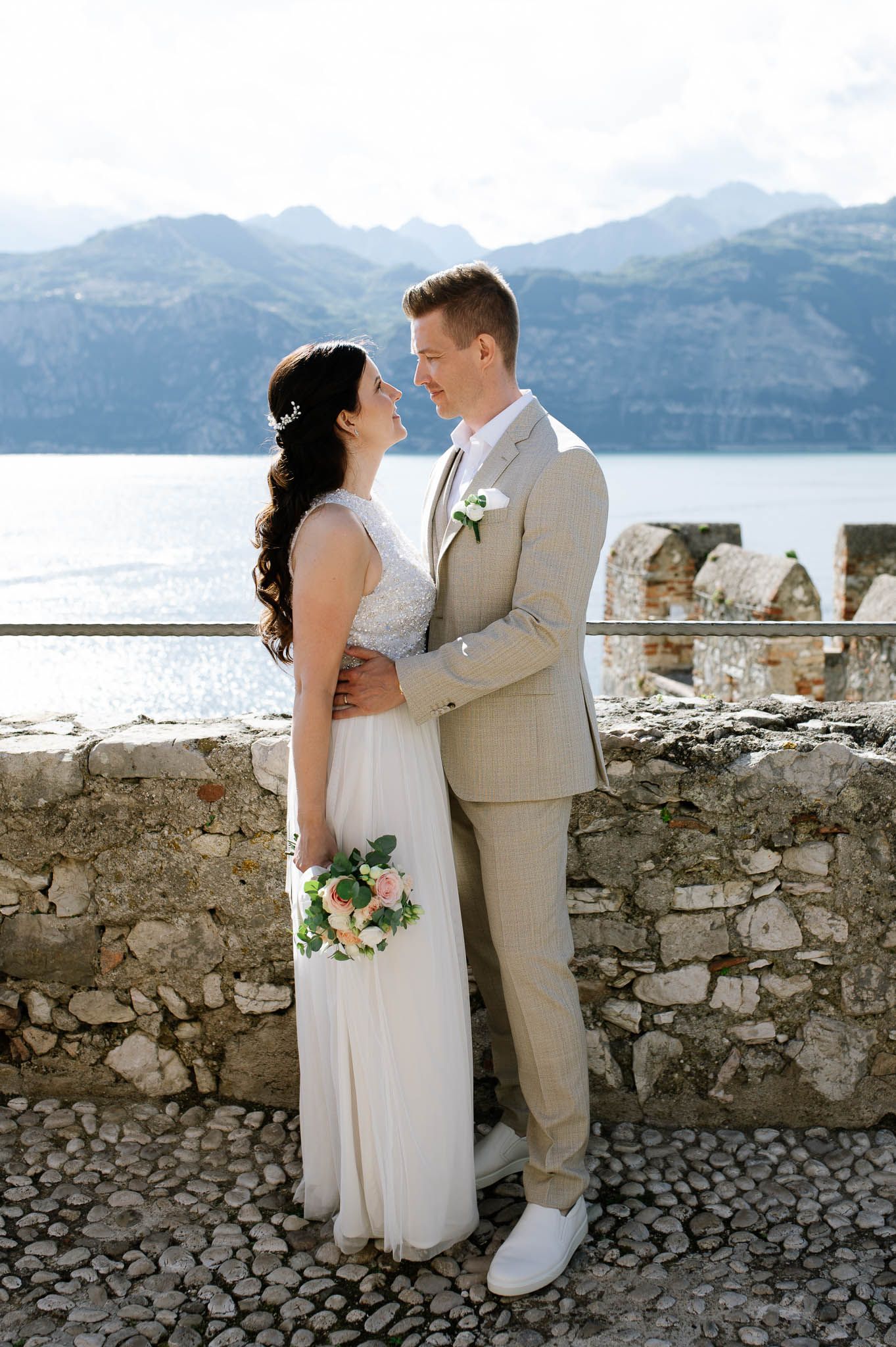 Bride and groom embrace on a stone terrace with stunning lake and mountain views behind them.