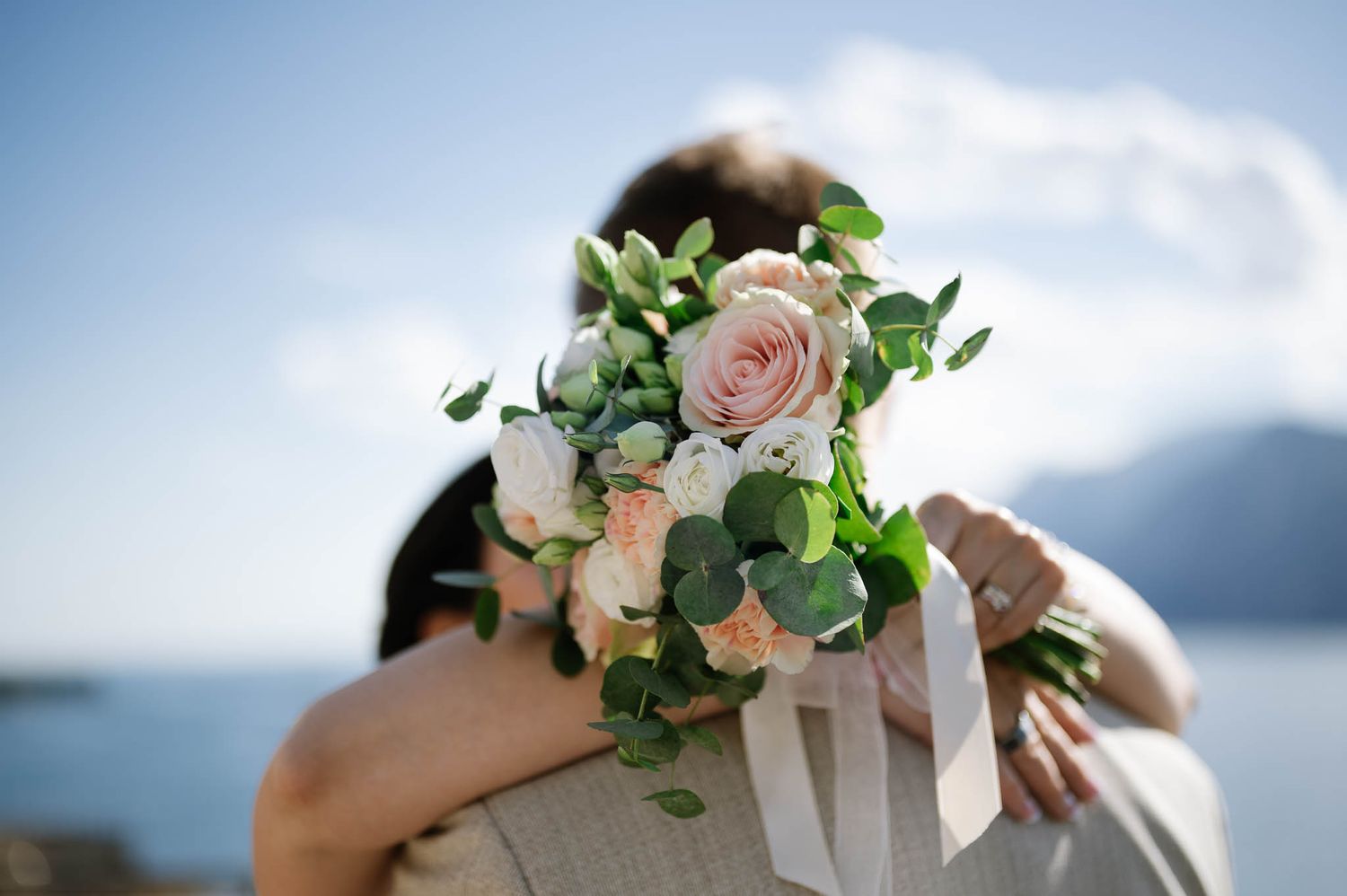 Close-up of a bridal bouquet with pink roses and greenery as couple embraces with lake backdrop.