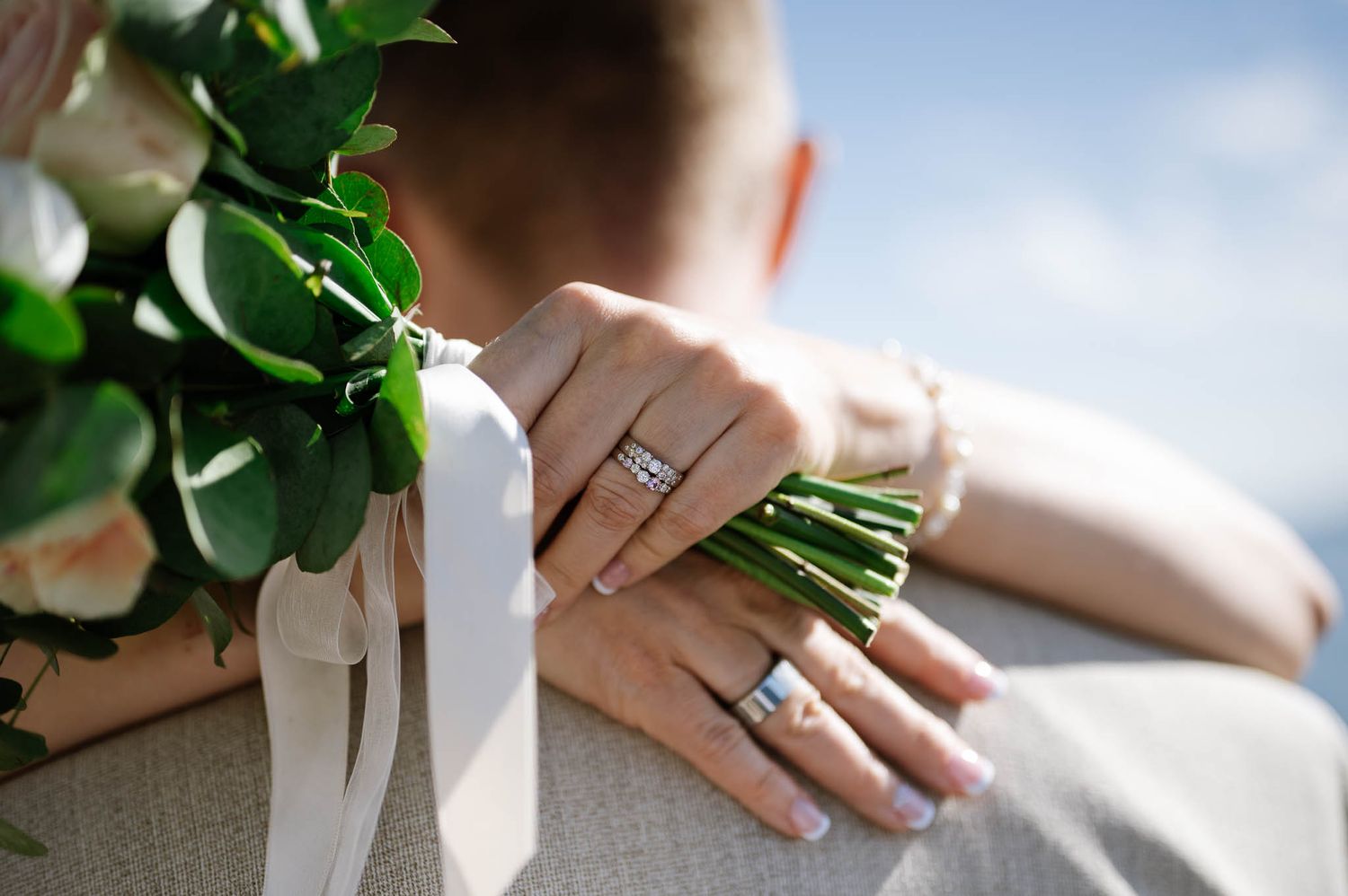 Close-up of bride and groom wedding rings on hands holding a white and pink bouquet.