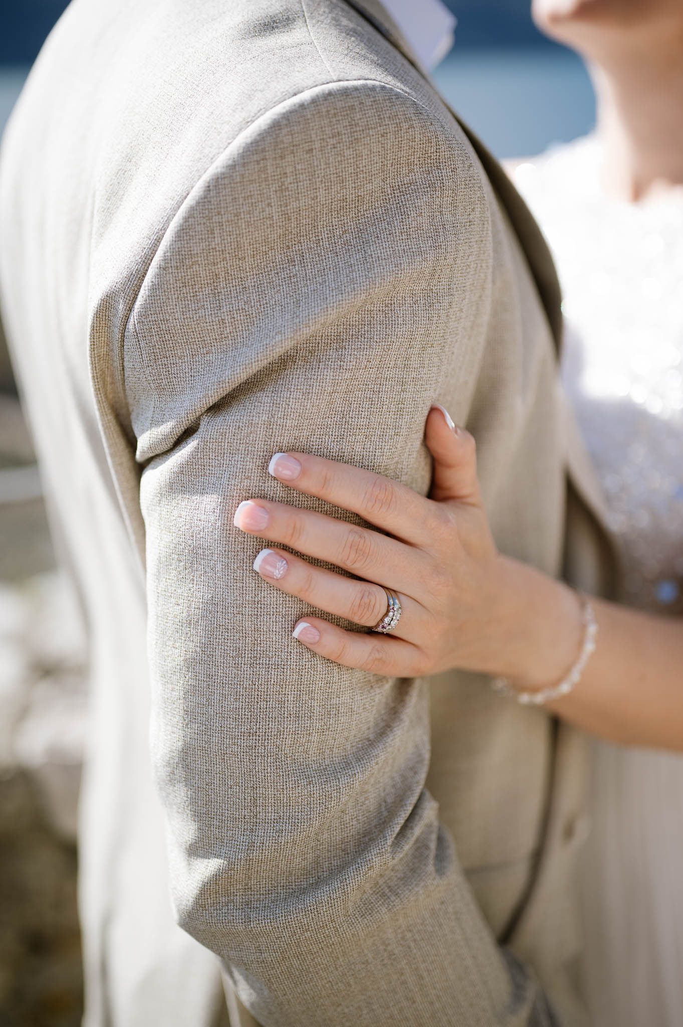 Close-up of a bride's hand with engagement ring resting on groom's gray suit jacket sleeve.