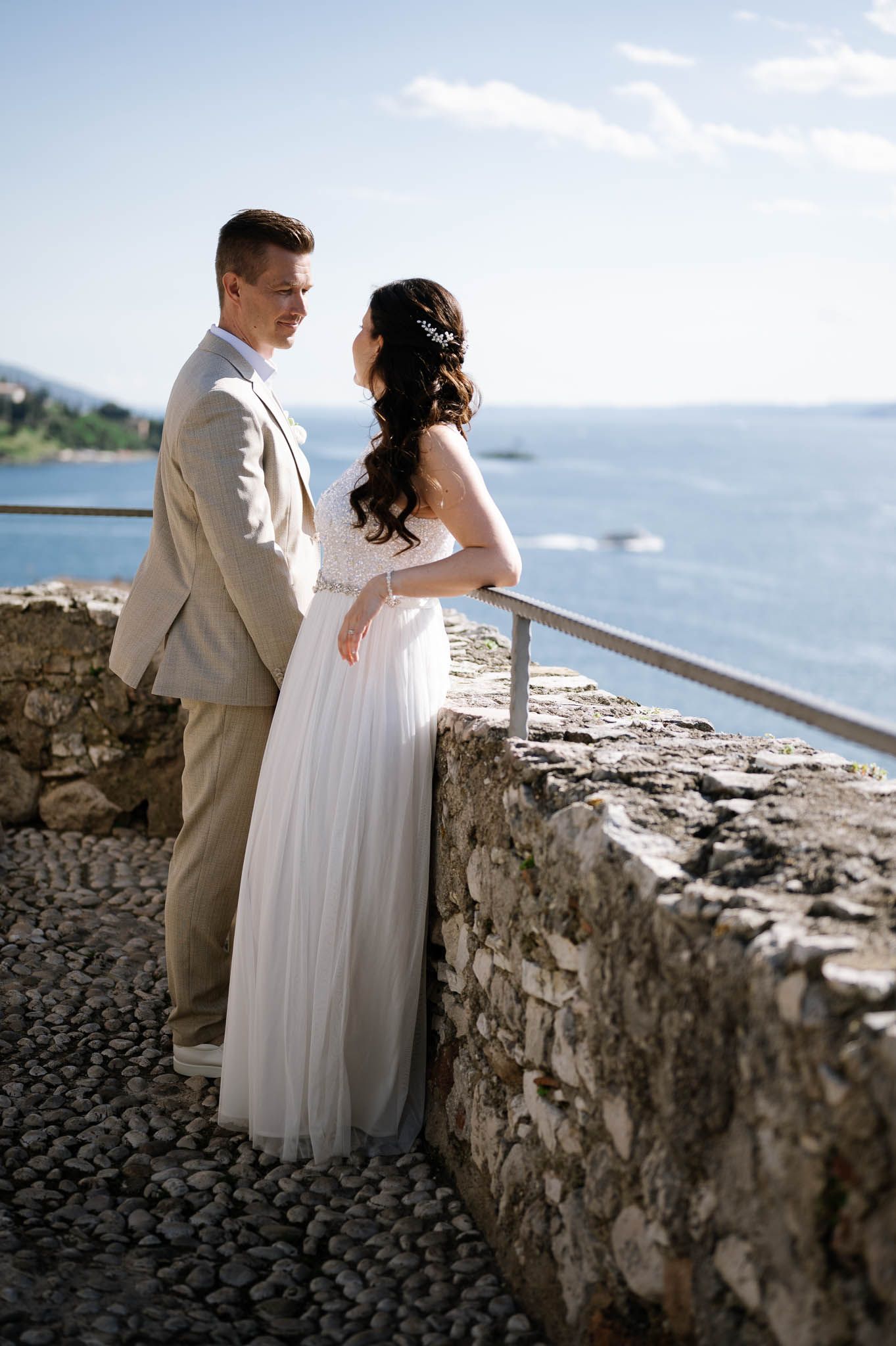 Bride in white tulle gown and groom in beige suit stand together by a stone wall with lake views.