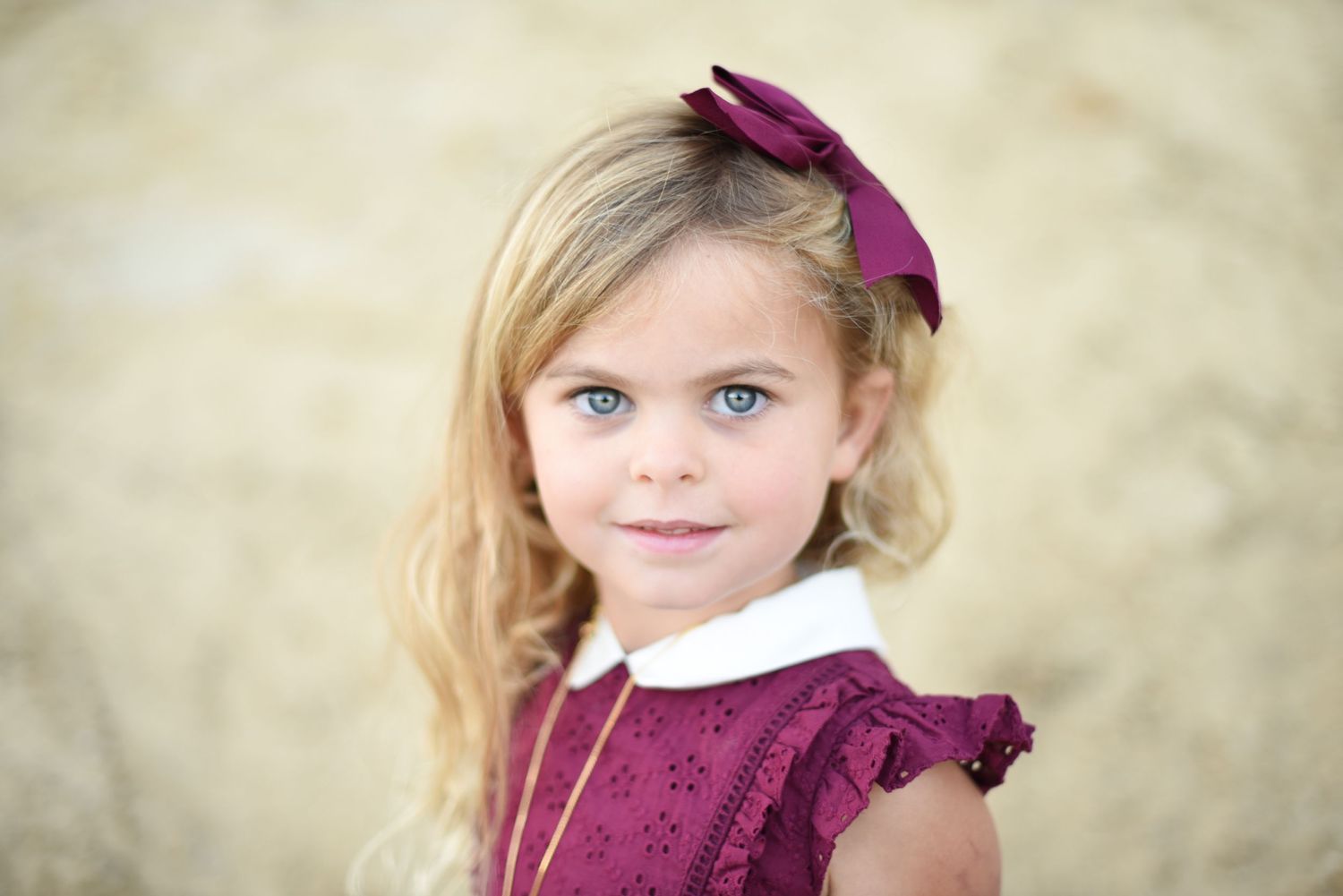 Young child in burgundy dress and bow posing sweetly against soft background.