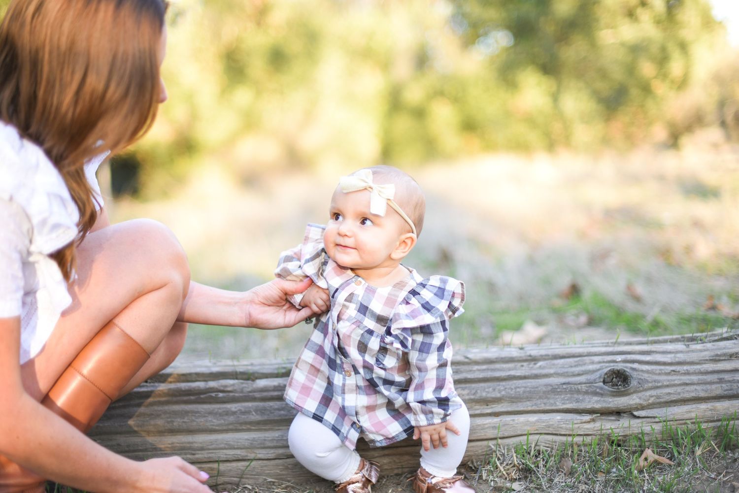 A small child in a plaid shirt sits on a rustic wooden log while interacting with someone outdoors.