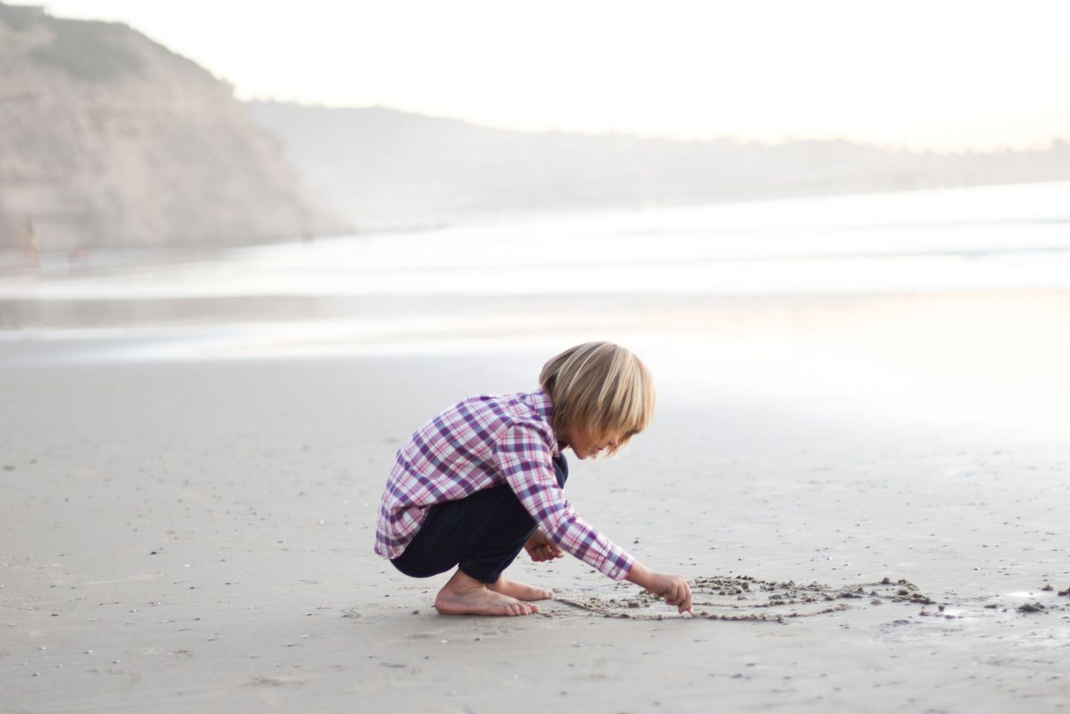 Child exploring and playing in the sand at a misty beach during sunset.