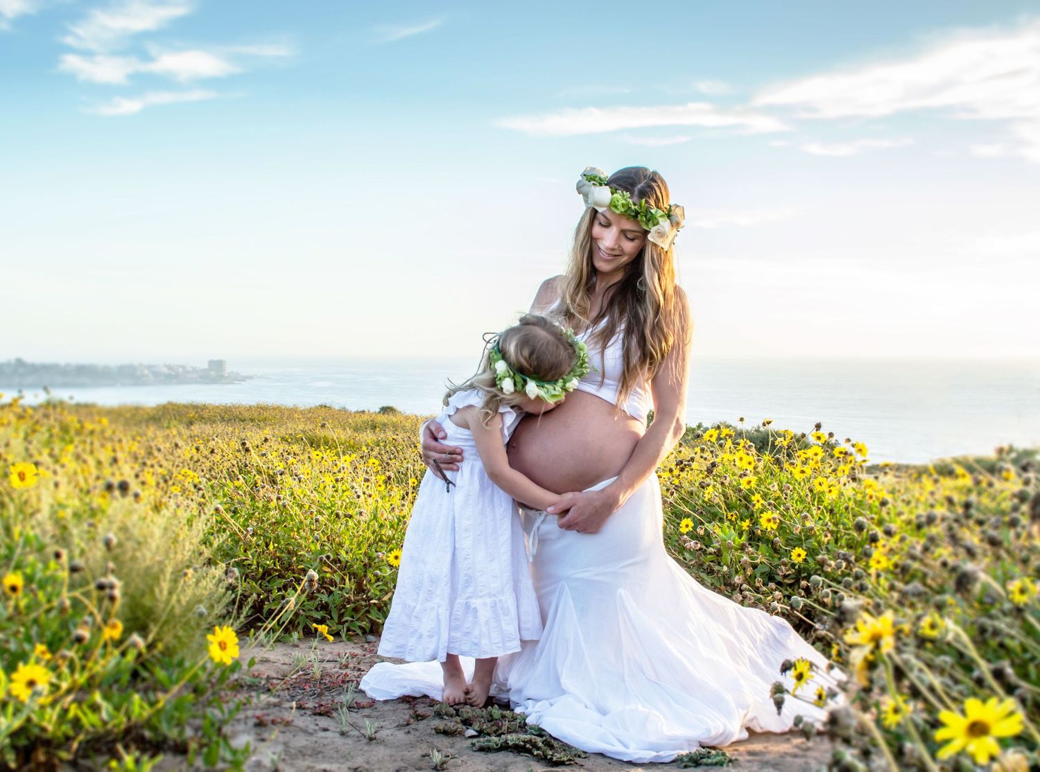 Expectant mother in white dress poses in field of yellow wildflowers against blue sky.