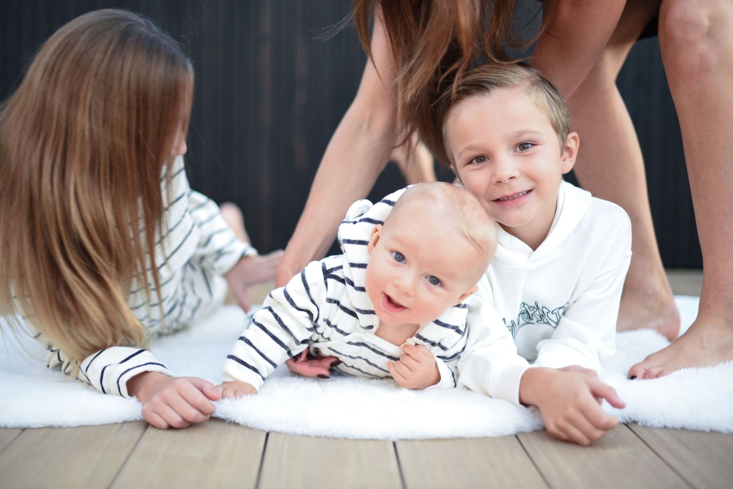 Three young siblings lying together on a white rug while wearing matching white and striped clothing. In home mini photoshoot, San Diego mom