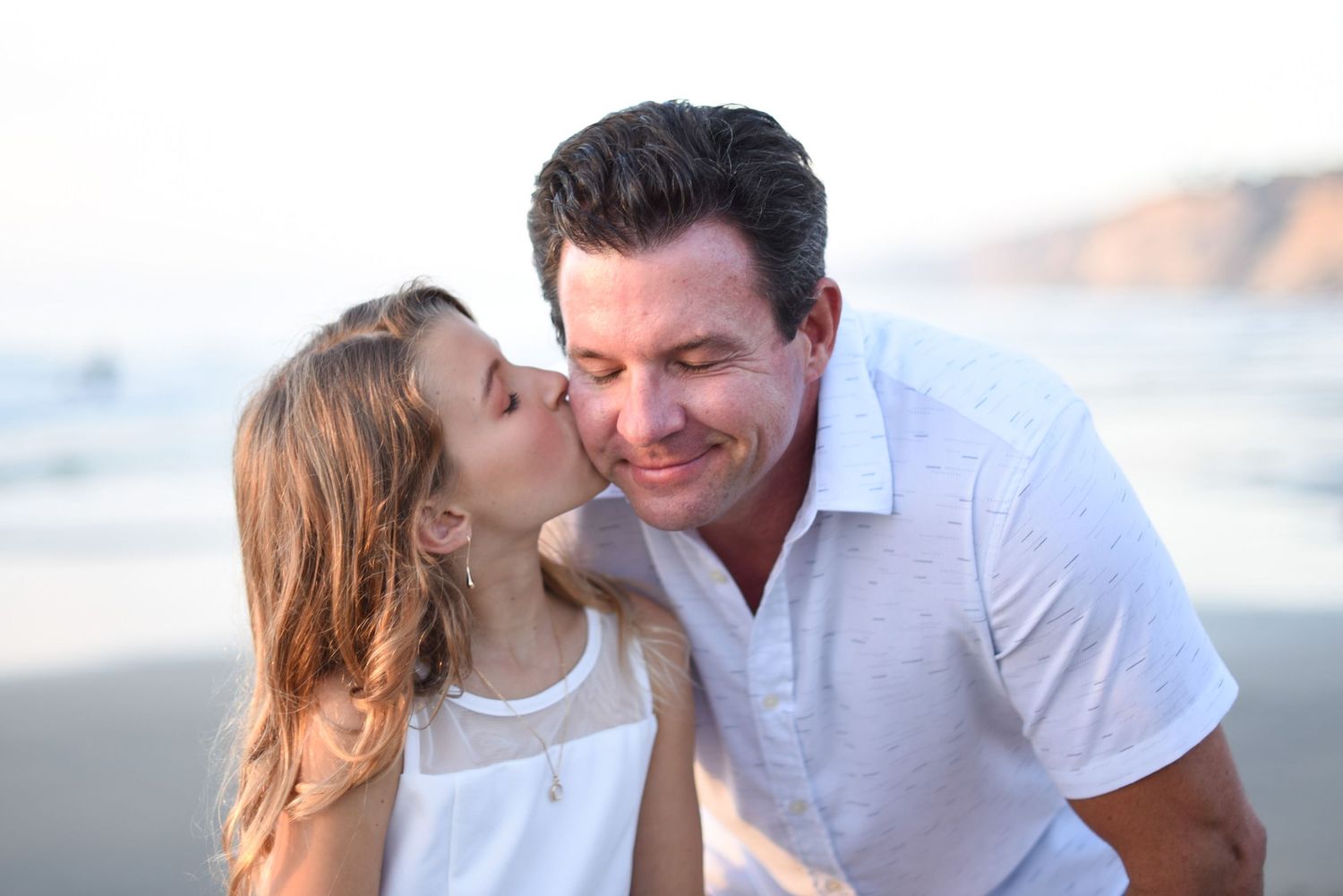 Father daughter moment. share a tender moment together on a sunny beach with ocean waves and cliffs in the background.