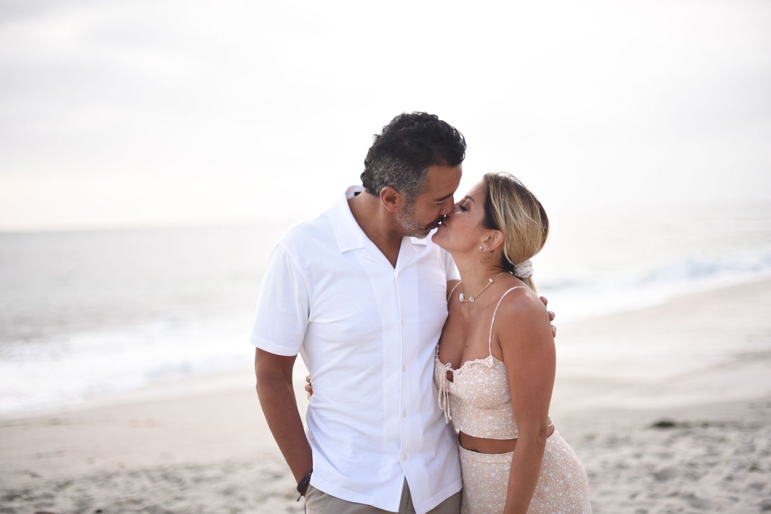 Romantic couple embracing on a beach in white outfits during sunset.
