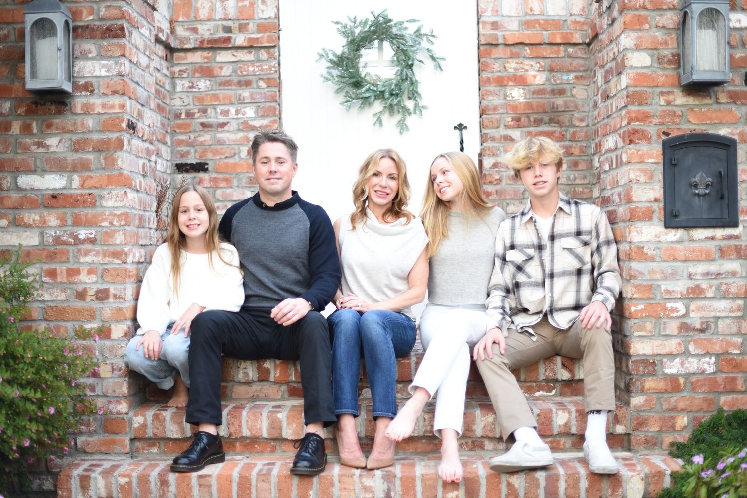 Family sitting on brick steps with wreath hanging on rustic wall.