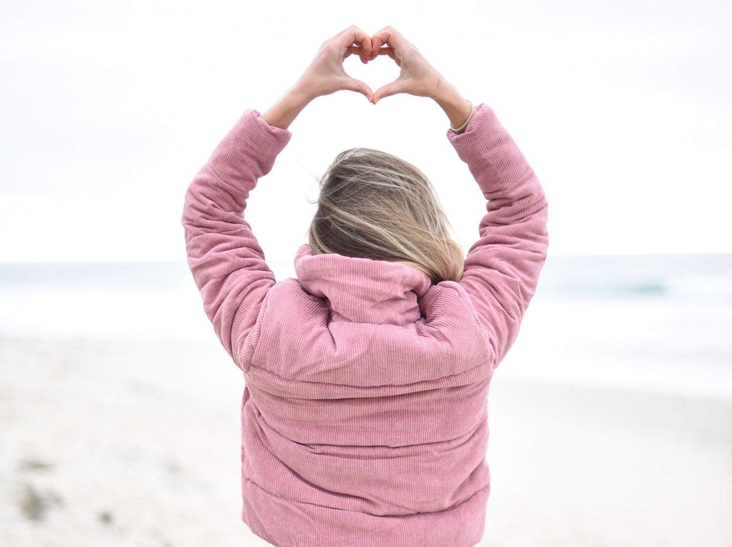 Person in pink sweater making heart shapes with hands against beach background.