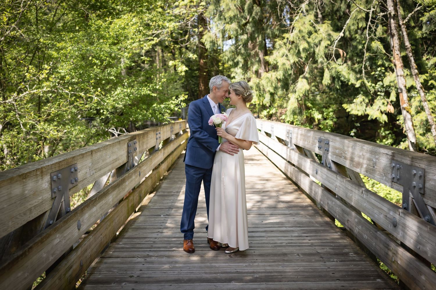 A couple shares a romantic kiss on a wooden bridge surrounded by lush green trees in an outdoor wedding setting.