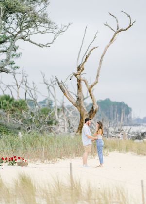 Happy newly engaged couple on Jekyll Island beach during surprise engagement picnic pictures with roses, champagne and proposal on Driftwood