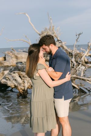 couple embracing during proposal of young couple on beach in south georgia's vacation destination jekyll driftwood trees behind them