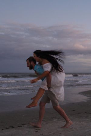 Woman wearing a white dress, man carrying woman on the beach during sunset engagement session Jekyll Island near Great Dunes, Corsair