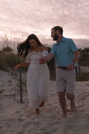Woman wearing a white dress, man wearing casual summer clothing, running on the beach on the sand sunset engagement session Jekyll Island
