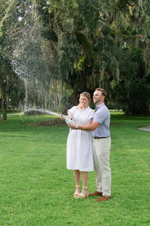 Couple popping champagne, woman wearing white dress, man wearing summer casual clothing on Jekyll Island during their engagement photos
