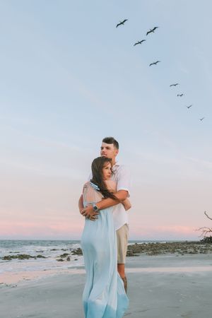 Couple holding each other after engagement on beach on Jekyll Island. Pelicans and beautiful sunset behind them