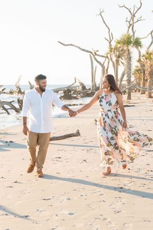 Couple holds hand and walks along beach during sunrise picnic mimosa proposal on Jekyll Island's driftwood beach.
