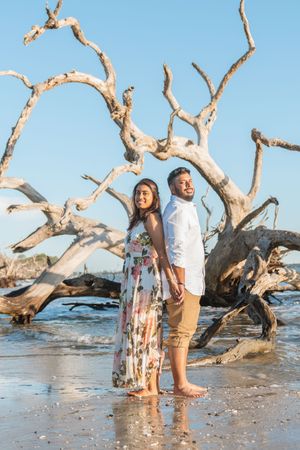 engagement portraits with driftwoods behind couple during a sunrise beach proposal, planned to surprise your bride to be