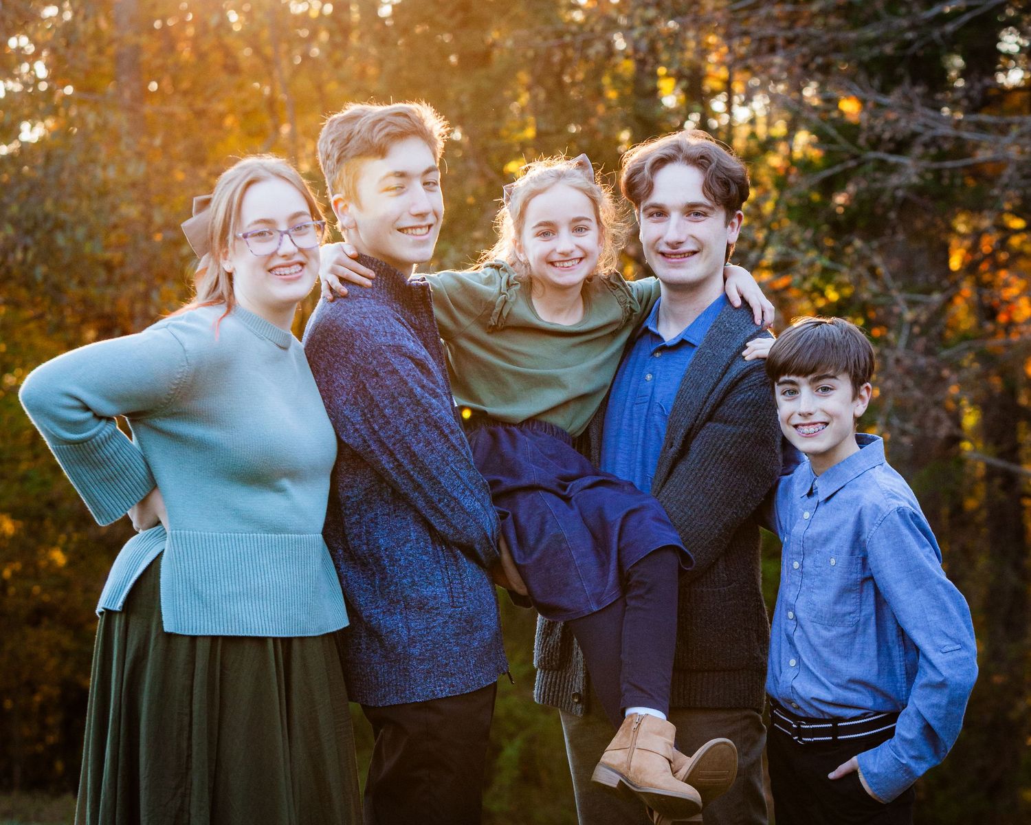Five siblings smiling together in a warm autumn outdoor setting with golden trees in the background.