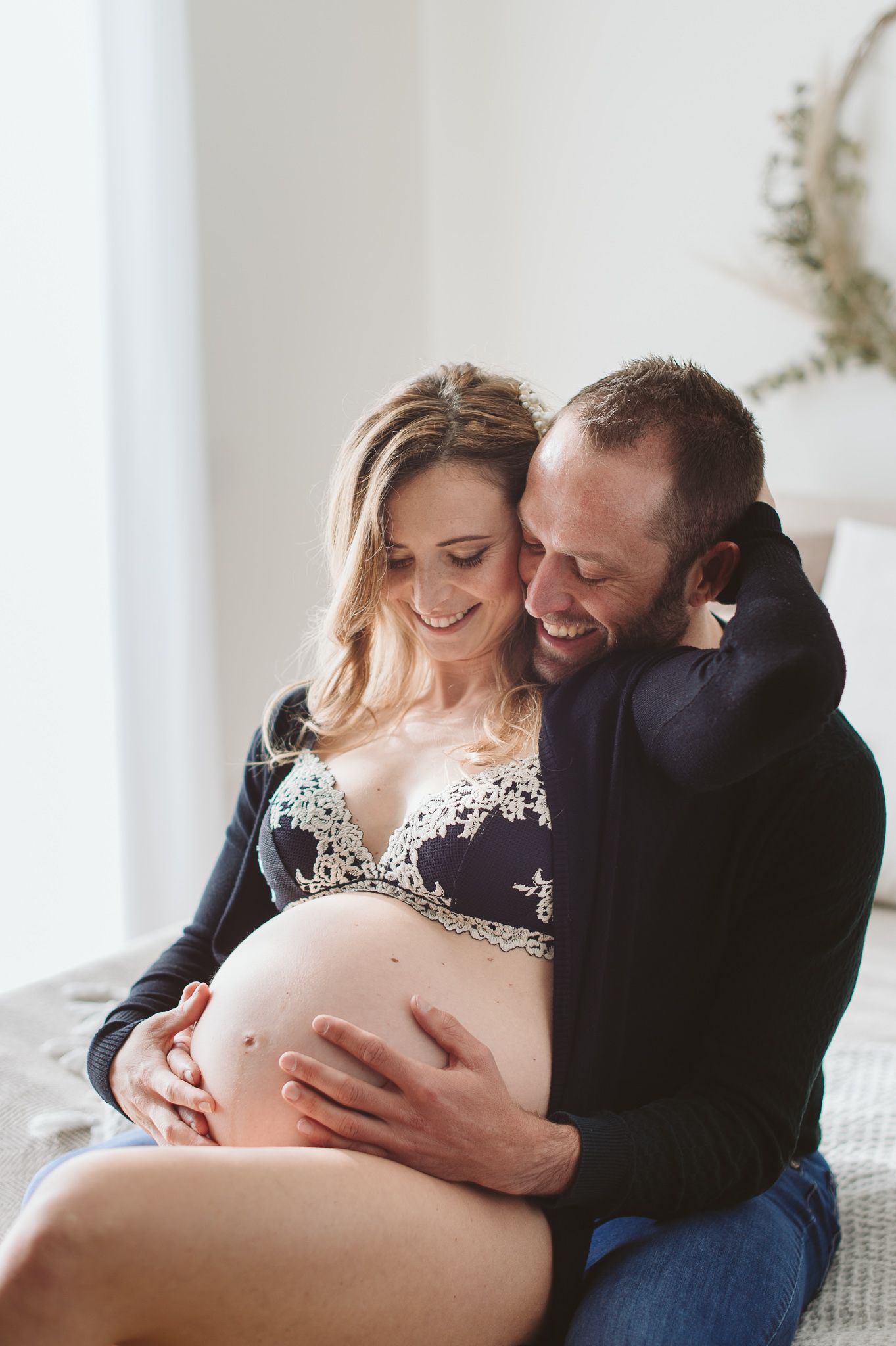 Happy couple sharing a tender maternity moment, cradling pregnant belly together while sitting on a bed.