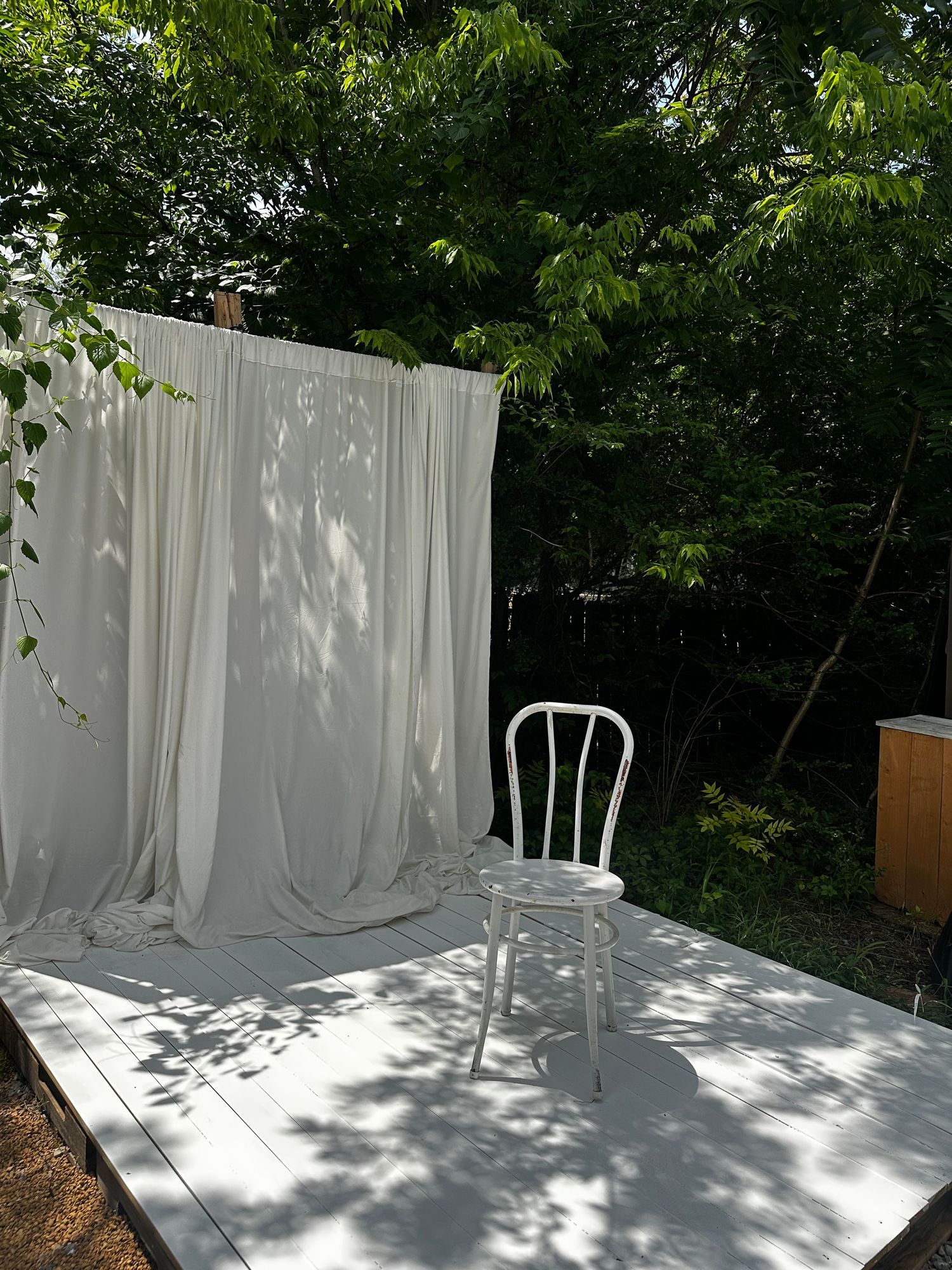 Outdoor photography setup with white draped backdrop, white chair on wooden platform, surrounded by lush green trees.