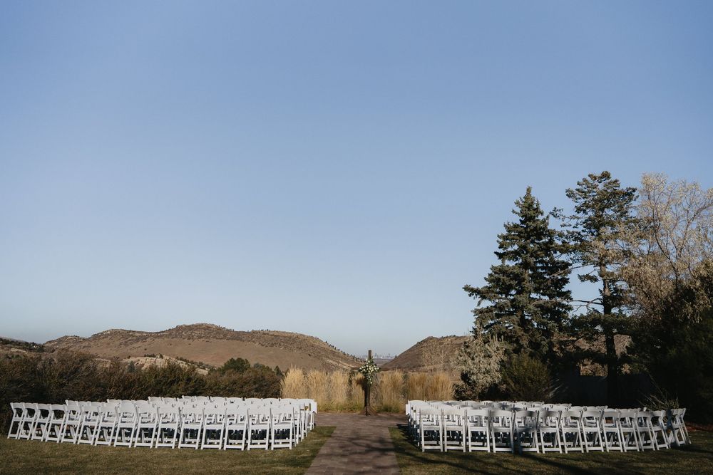 Outdoor wedding ceremony setup with white chairs arranged in rows, surrounded by trees and mountain backdrop under clear blue sky.