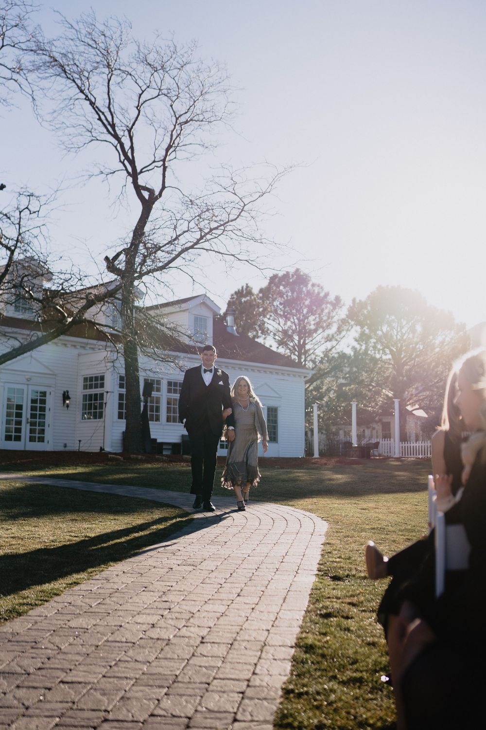 Couple walks down brick path toward white estate venue on sunny winter wedding day.