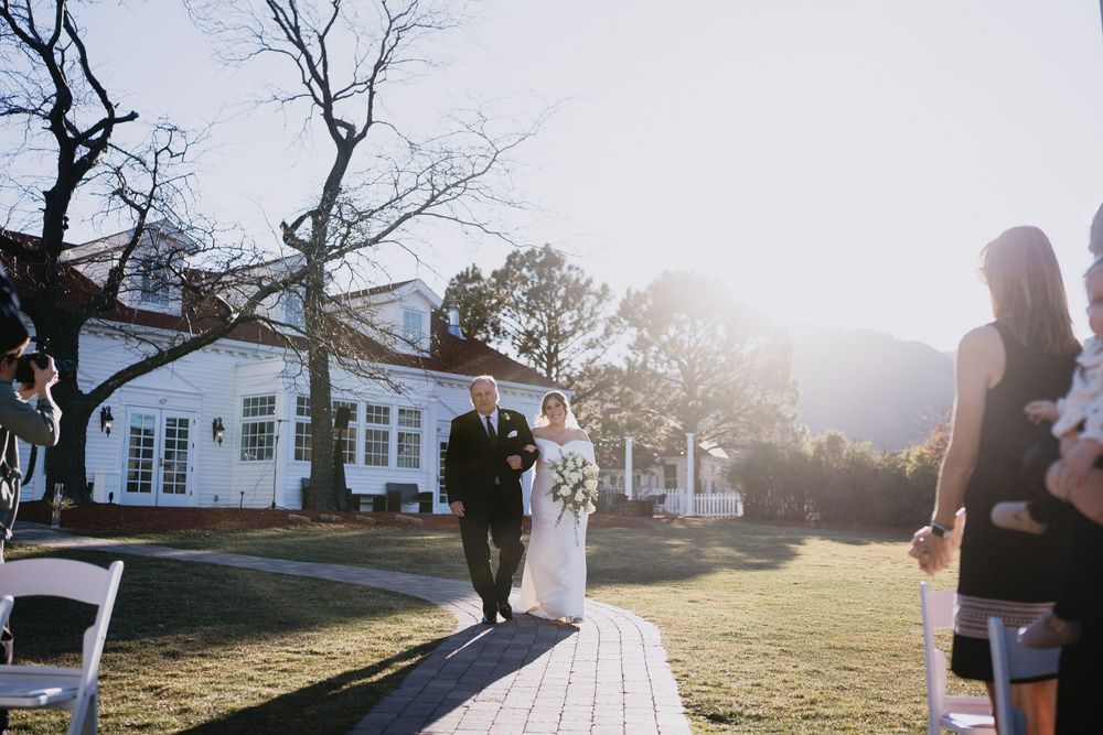 Bride and groom walk down outdoor aisle at elegant white estate wedding venue with mountain backdrop.