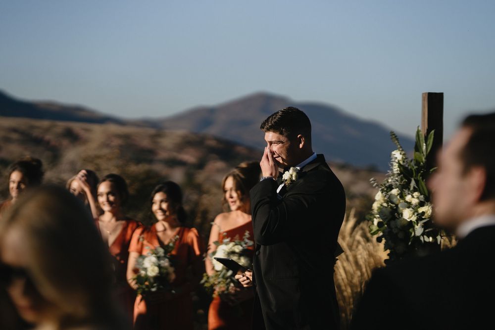 Emotional groom wiping tears at outdoor wedding ceremony with bridesmaids in rust dresses and mountain backdrop.