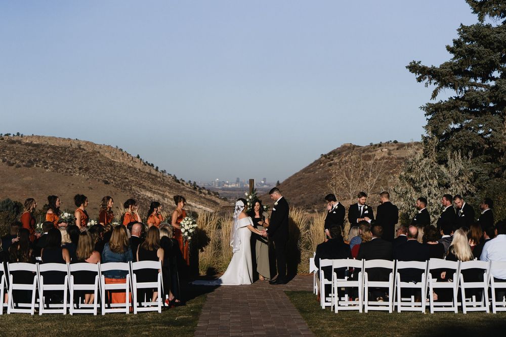 Outdoor wedding ceremony with bridal party in black and orange attire, white chairs, and scenic hillside backdrop under clear blue sky.
