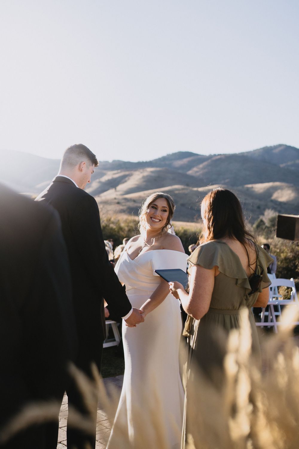 Bride smiling joyfully during outdoor wedding ceremony with mountain backdrop, wearing off-shoulder white gown.