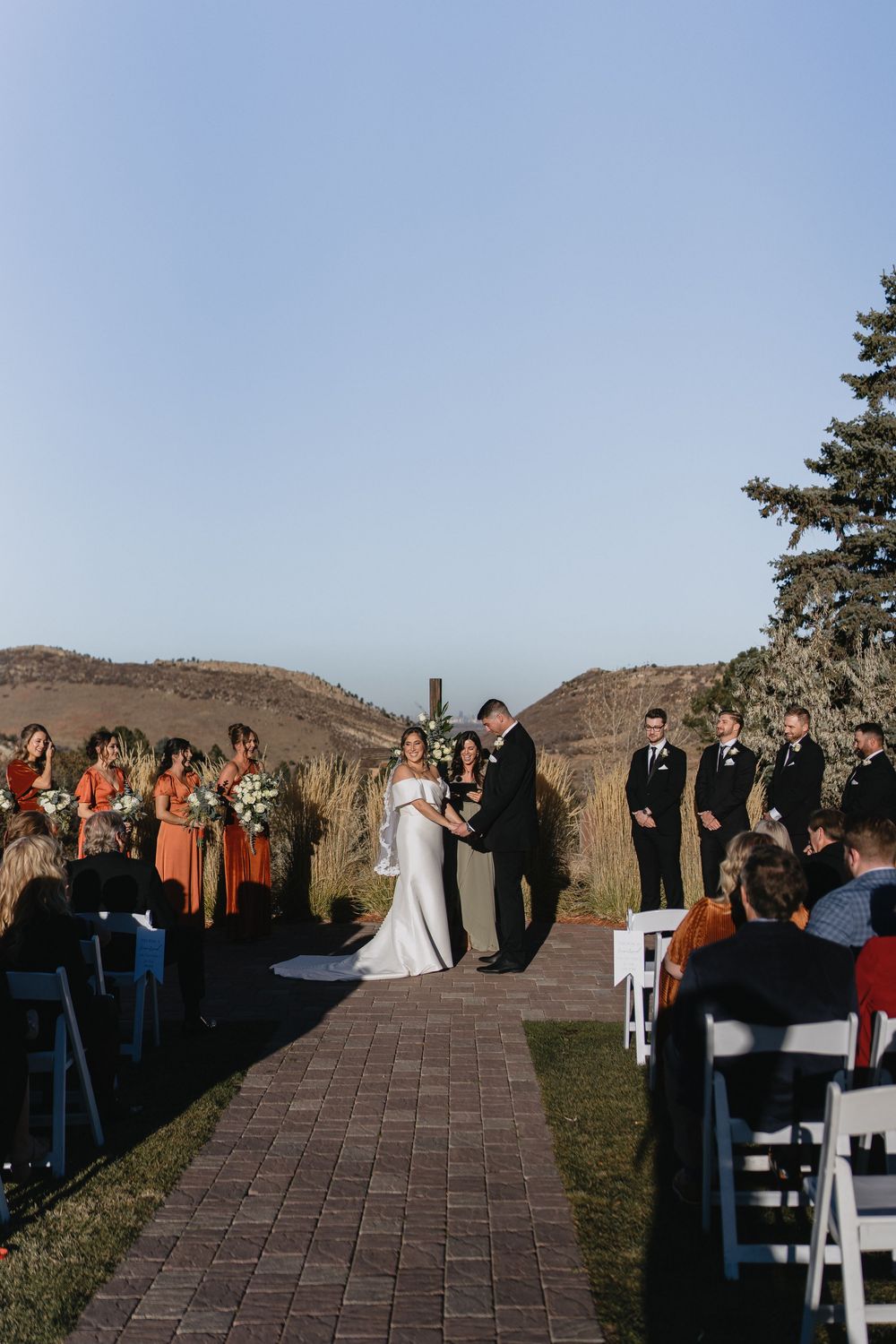 Outdoor wedding ceremony with bride and groom at altar, bridesmaids in orange dresses, groomsmen, and guests seated on white chairs.